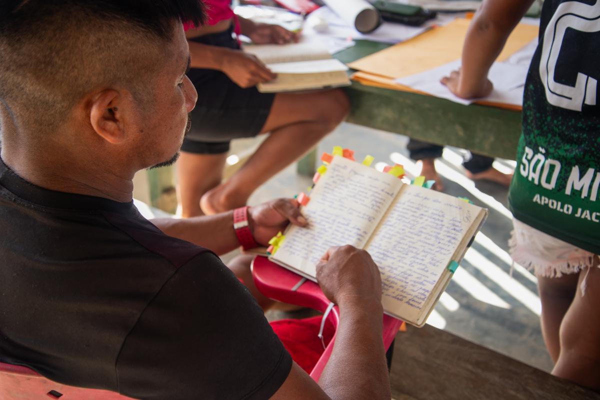 Homem aparece na foto sentado de costas e com um caderno com registros dos AIMAs nas mãos